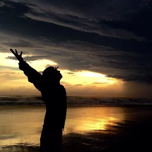Silhouette of Raven Indigo Carrigg standing at the ocean with arms open beneath a dramatic sky.